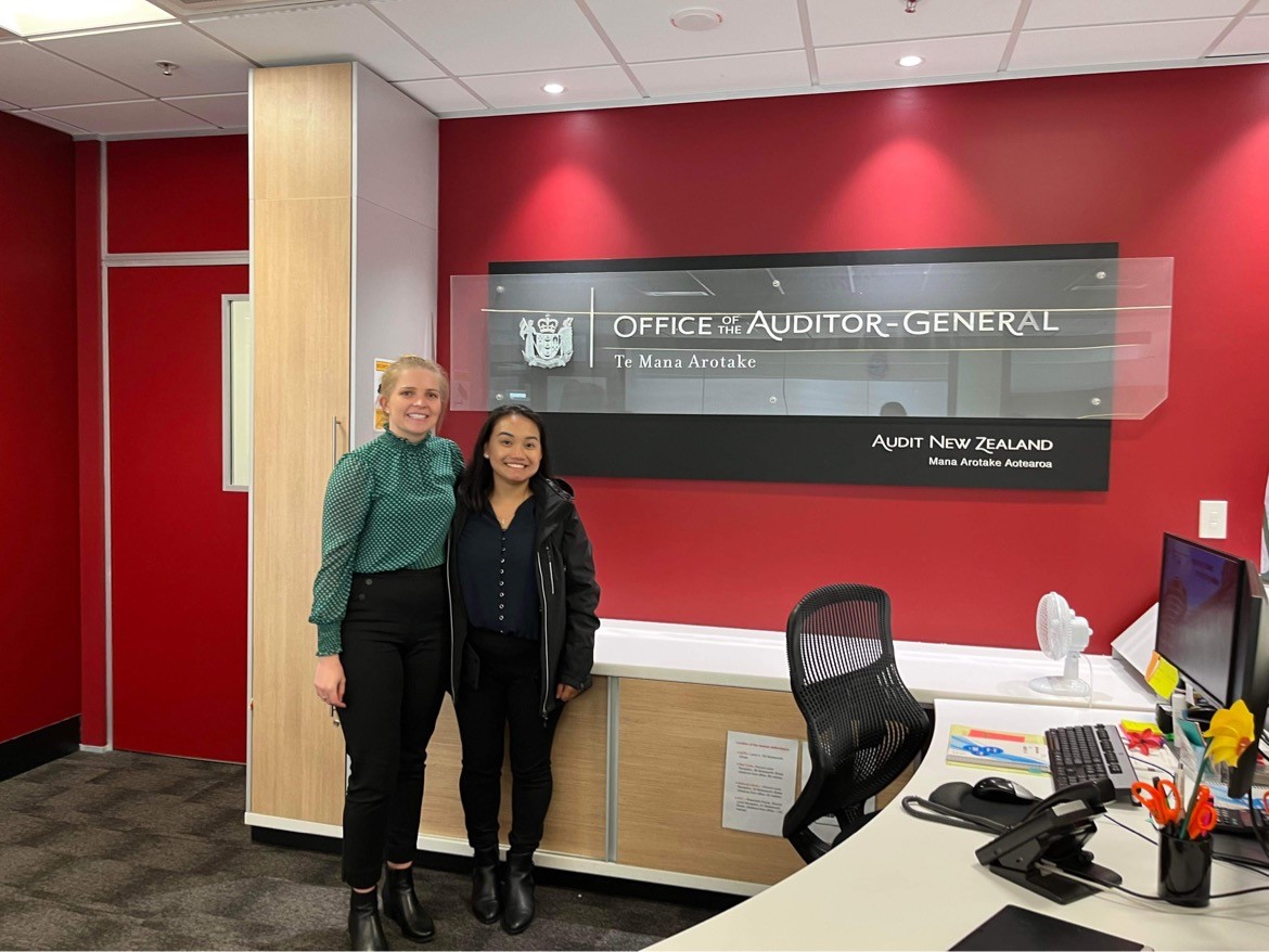 Image of Eliza and Khendie standing in front of the Audit New Zealand sign in the office.
