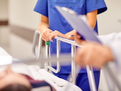 Image of part of a hospital bed, with medical personnel standing beside it and a patient chart 