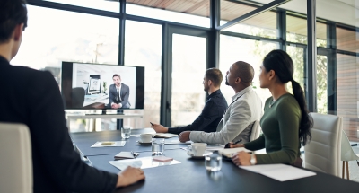 Four people sitting around an meeting table, looking at one person on a screen.