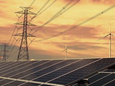 Foreground show solar panels on the roof of a building. Power lines can be seen into the distance. Wind turbines are in the background. 